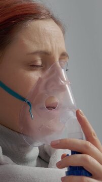 Vertical Screen: A woman with a mask inhales steam from a saline solution to treat her cough. She uses a nebulizer for her respiratory infection during the day at home.