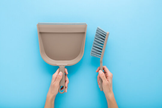 Young adult woman hands holding and showing new brown broom and dustpan for home floor sweeping on light blue table background. Pastel color. Closeup. Point of view shot. Flat lay. Top down view.