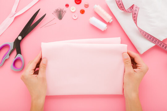 Woman hands holding folded light pink gabardine elastane fabric for female clothes making. Sewing items on pink table background. Pastel color. Closeup. Flat lay. Point of view shot. Top down view.