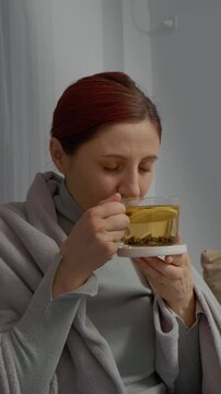 Vertical Screen: A woman sits on the couch holding a cup of herbal tea. She uses napkins while trying to feel better from her cold. She hopes to recover from her illness soon.