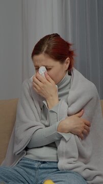 Vertical Screen: A woman sits on the couch wrapped in a blanket. She sneezes and coughs as she sips herbal tea to feel better while struggling with cold symptoms.