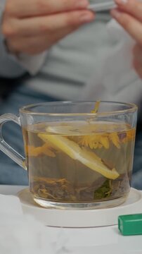 Vertical Screen: A woman sits in bed, checking her temperature while sipping herbal tea. She is dealing with a cold and tries to recover from her symptoms and fever.