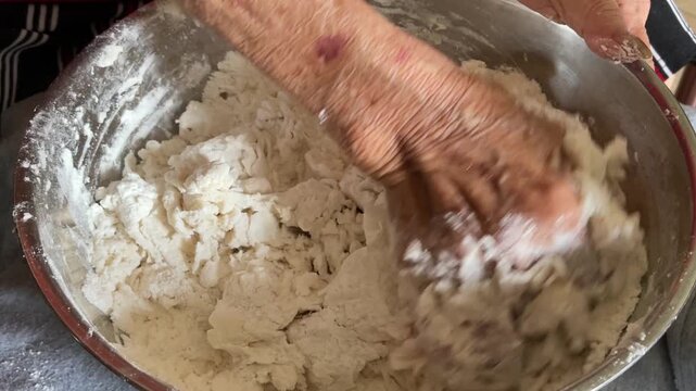 An elderly person&rsquo;s hand are shown in a close-up, continuously kneading a mixture of flour and water in a metal bowl to form a solid dough for traditional buns.