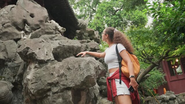Young woman exploring rock garden, reaching toward weathered scholar rock, wearing backpack and red jacket tied at waist, curly hair pulled back, hand touching porous stone, wooden pavilion and dense