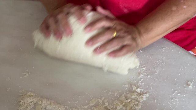 A close-up shot captures an elderly person's hands vigorously kneading a smooth white dough on a white table to prepare traditional handmade baozi.