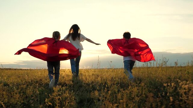 superhero girl boy mother play green grass. concept cute happy family being green field sunset. family winners together friend. superhero concept. happy superhero family jogging outdoors red raincoat.