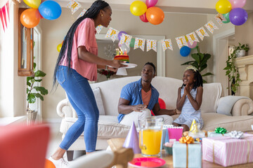 Naklejka premium African American family holding birthday party in living room, woman carrying cake with lit candles