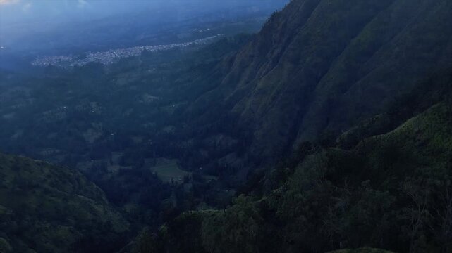 Aerial view of a deep mountain valley at dusk, with a small town&rsquo;s lights in the distance.