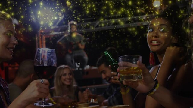 Friends at table raising wine, whisky glasses, clapping to live music cue, gold sparkles drifting