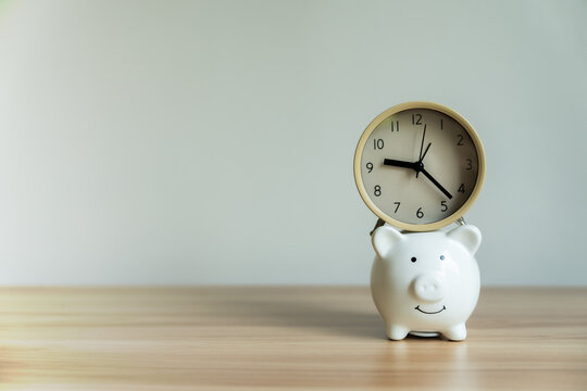 White piggy bank smiles gently on wooden table. Clock rests atop it, hands point to time. Minimalist scene suggests budgeting or savings. Clean background offers copy space for text