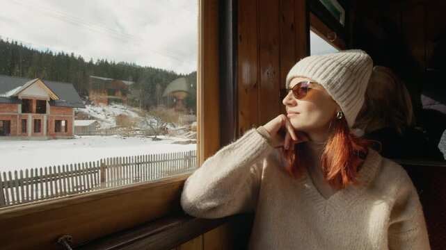 A woman rides on an old train in Romania. She looks out the window and enjoys the views of nature as the train travels along its path through the landscape.