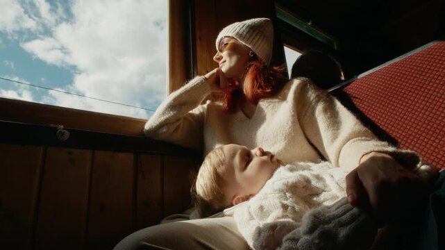 A mother holds her sleeping boy while riding on an old train in Romania. She looks out the window, enjoying the sights during their journey.