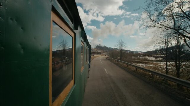 The Mocanita steam train moves along the path in Bucovina, Romania, passing through small villages and releasing thick smoke into the air as it travels.