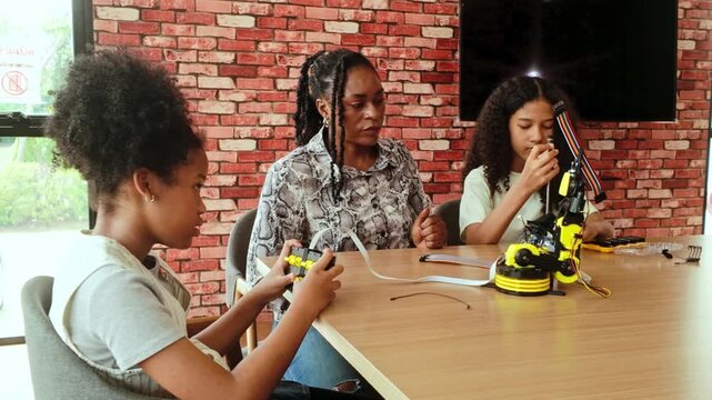 Instructor guides two girls building a yellow robotic arm during a STEM workshop at a table with tools, in a room with a brick wall