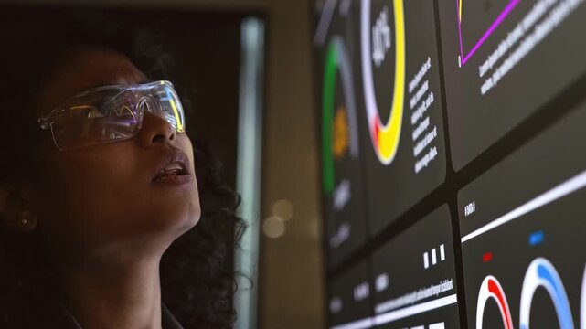 Person wearing safety glasses examines colorful data dashboards on large monitors in a dim tech workspace.