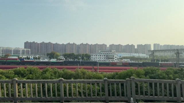 Aerial view from elevated train passing Suzhou freight rail yard in spring, with shipping containers and residential towers in background.