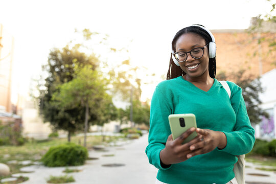 Smiling student walking and using smartphone with headphones in the park