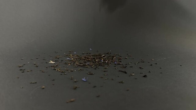 Dried earl grey tea leaves with blue cornflower petals falling in slow motion onto a dark surface, creating a small pile of aromatic organic bergamot blend for brewing