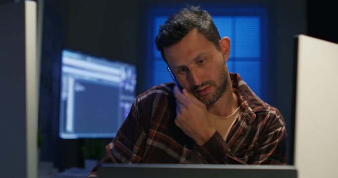 The programmer working while talking telephone .He working late with multiple task with overworked at night time.