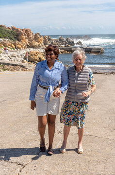 Western Cape South Africa. 05.11.2026. .Two women of different ages and ethnicities share a moment of connection while walking together on a sunny day by the sea, with crashing waves in the background
