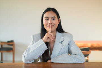 Smiling Thai woman wearing a blazer making shush gesture at a table, capturing a natural,...