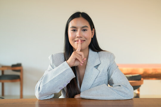 Smiling Thai woman wearing a blazer making shush gesture at a table, capturing a natural, professional lifestyle moment of a happy Isan student.
