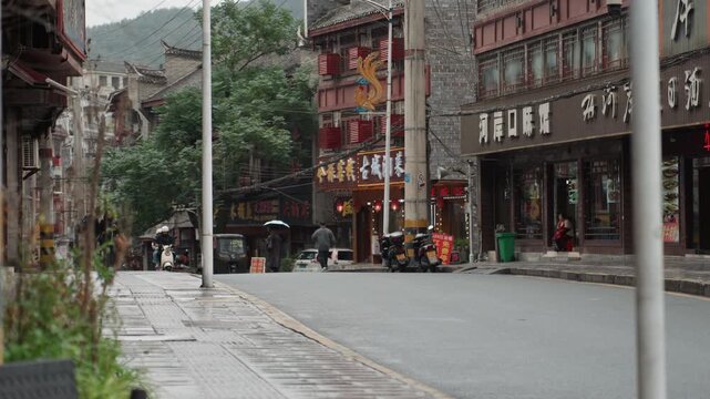 Quiet wet street with storefronts, narrow sidewalk, parked scooter, delivery rider approaching, muted signage, overcast sky, distant treecovered hill, subtle puddles reflecting neon, slowpaced morning