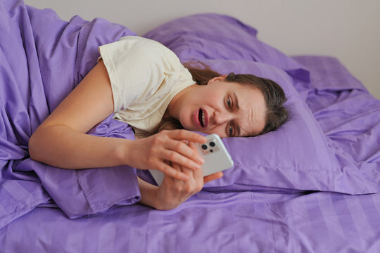 A person is lying in bed under purple covers while looking at a phone with surprise. The setting is a bedroom during daytime with soft light