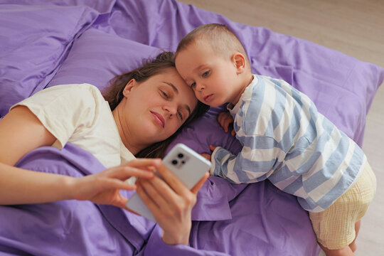 A mother and her young child are lying on a bed with purple bedding. They are looking at a smartphone together, sharing a moment during morning time