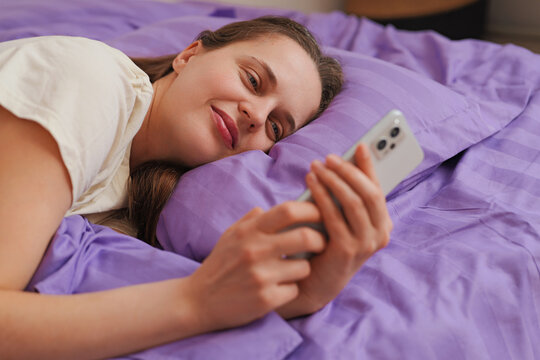 A woman lies on a bed with purple sheets, looking at her smartphone and smiling. It is afternoon and the room is warm and inviting as she enjoys her time