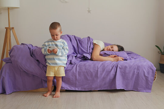A child is standing beside a bed while a mother lays asleep on a purple blanket. The room is simple with a lamp and some plants in the background
