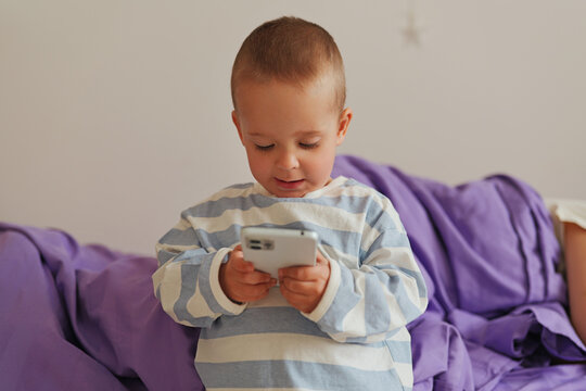 A child sits on a bed and looks at a smartphone. The room has soft light coming in. The child seems focused on the device in their hands