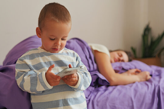 A child stands next to a bed, focused on a smartphone. An adult sleeps peacefully under a blanket. Morning light fills the quiet room