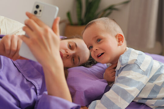 A child smiles as he leans close to his mother who looks at her phone. They lay on a bed with purple sheets in a bright room, enjoying their time together