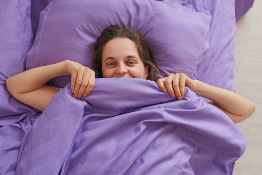 A woman is lying in bed with purple sheets. She is smiling and holding the blanket close as she enjoys a relaxed morning at her home
