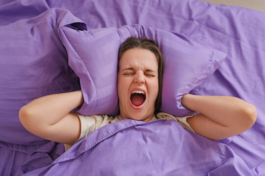 The person sits in bed, covering their ears while yawning. Purple sheets cover the bed. It appears to be morning time as they look tired