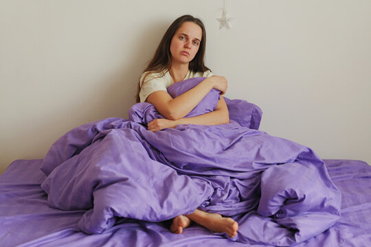 A woman sits on a bed with a purple blanket wrapped around her. She looks thoughtful and is resting her chin on her knees. The room is bright