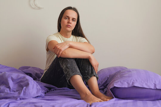 A woman sits on a bed with purple bedding in a simple room. She looks directly at the camera with a serious expression. Sunlight enters through the window