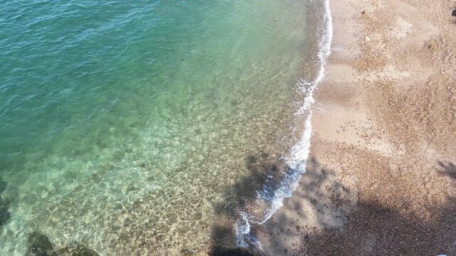 crystal clear turquoise sea water with small waves gently lapping onto a sandy pebble beach. Natural summer sunlight reflecting on the water surface. Refreshing vacation background.	
