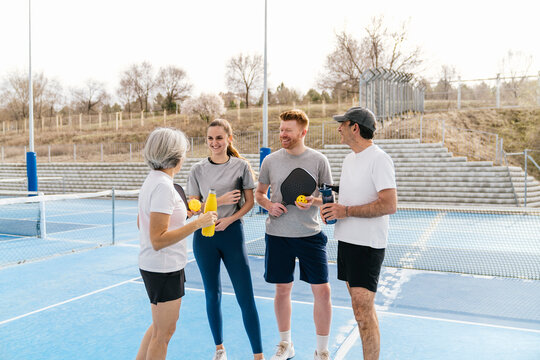 Two senior adults and two young adults enjoying a break from playing pickleball on an outdoor court, holding racquets and drinks
