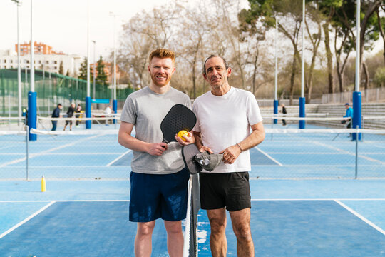 Two smiling men standing on a blue pickleball court, holding paddles and ball, ready for play outdoors