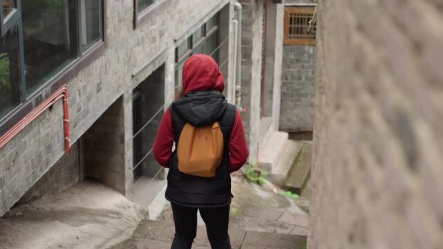 Red hoodie backpack navigating narrow concrete alley, lone urban explorer descends between brick buildings and metal railings, gritty textured walls, candid city atmosphere, muted tones and curious