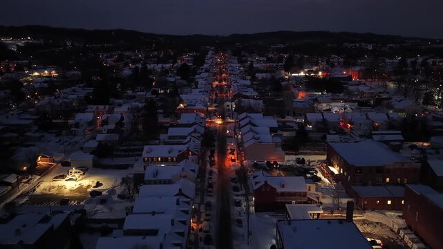 Slow drone flight over illuminated cozy Main Street of small American town in winter. Snowy roofs of row homes along road. Aerial flyover shot. Christmas season at night scene in USA. Peaceful city.