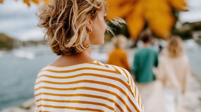 A woman in a striped top walking towards a group of people by the water, surrounded by stunning autumn foliage, portraying the beauty of nature and companionship.