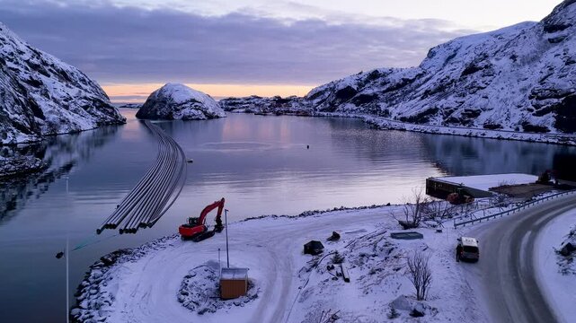 Cinematic aerial drone shot of snowcovered mountains surrounding a calm fjord in Lofoten, Norway during winter twilight. Features industrial pipes in water and a coastal road.