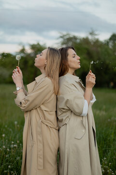 Two women blowing dandelions back to back in park wearing coats