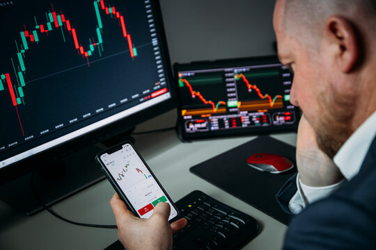 Stressfull trader holding smartphone, using trading app with stock market charts for financial analysis and monitoring investment data.