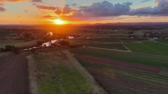 Aerial video flying over green and cultivated fields towards river and colourful sunset over Morpeth in NSW, Australia. Scenic rural landscape showing farmland patterns and waterway 