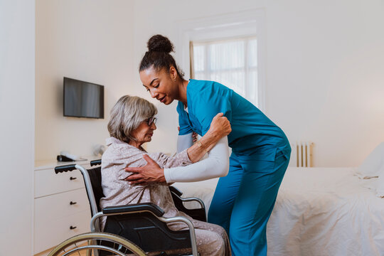 Caregiver helping senior woman transfer from wheelchair to bed at home
