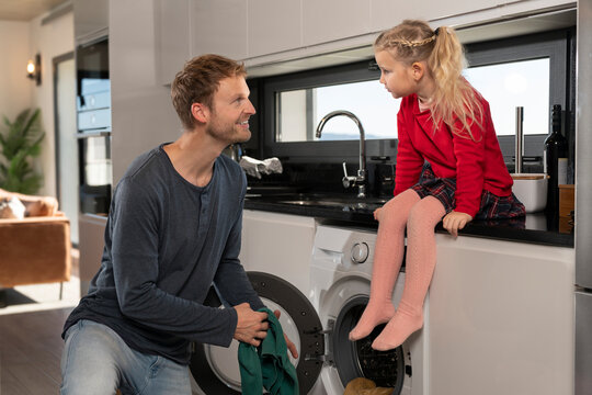 Father loading clothes in washing machine and talking with daughter siting on kitchen counter at home
